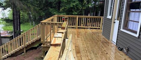 Una terraza de madera con una escalera de madera que conduce a otra terraza, rodeada de árboles y una casa con una puerta y ventanas blancas.