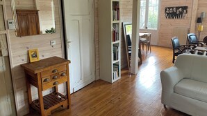 Una habitación acogedora con pisos de madera, un sofá blanco, una mesita de madera, una estantería y un comedor con sillas y una mesa.