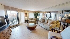 A spacious living room with a sofa set, a coffee table, and a TV. There are large windows with curtains, a plant, and a mirror. - Inlet Reef 413 2 Bedroom Condo by RedAwning (Destin)