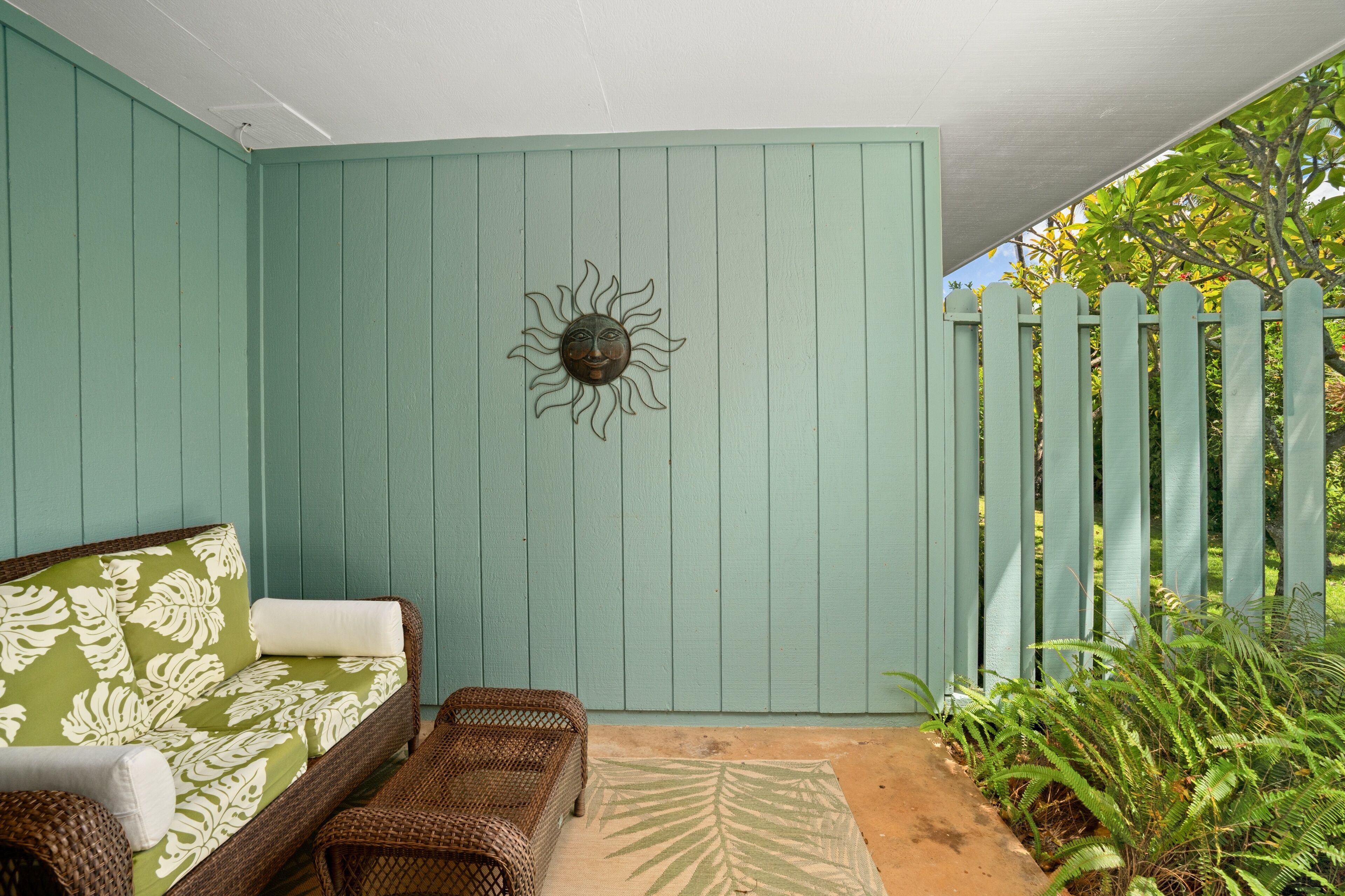 Un patio con pared verde, un sofá de mimbre con cojín estampado, una decoración de pared con forma de sol y una valla de madera con plantas.