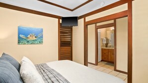 Un dormitorio con cama, almohadas, un cuadro de tortuga marina, televisión y un baño visible a través de una puerta abierta.