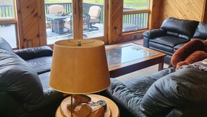 A cozy cabin interior with a leather sofa, a wooden coffee table, and a lamp. There are framed pictures on the walls and a view of a deck and trees outside. - Beautiful Lodge for Weddings, Reunions, Events or Family Gatherings. Sleeps 25+ (Redfield)