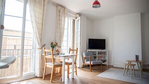 Un salon lumineux avec une table à manger, des chaises, un téléviseur et un balcon agrémenté de rideaux.