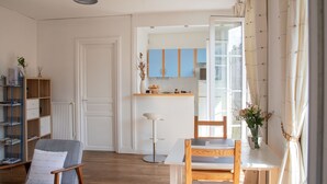 Un salon moderne comprenant un coin repas, une bibliothèque, un radiateur et une cuisine visible à travers une porte ouverte.