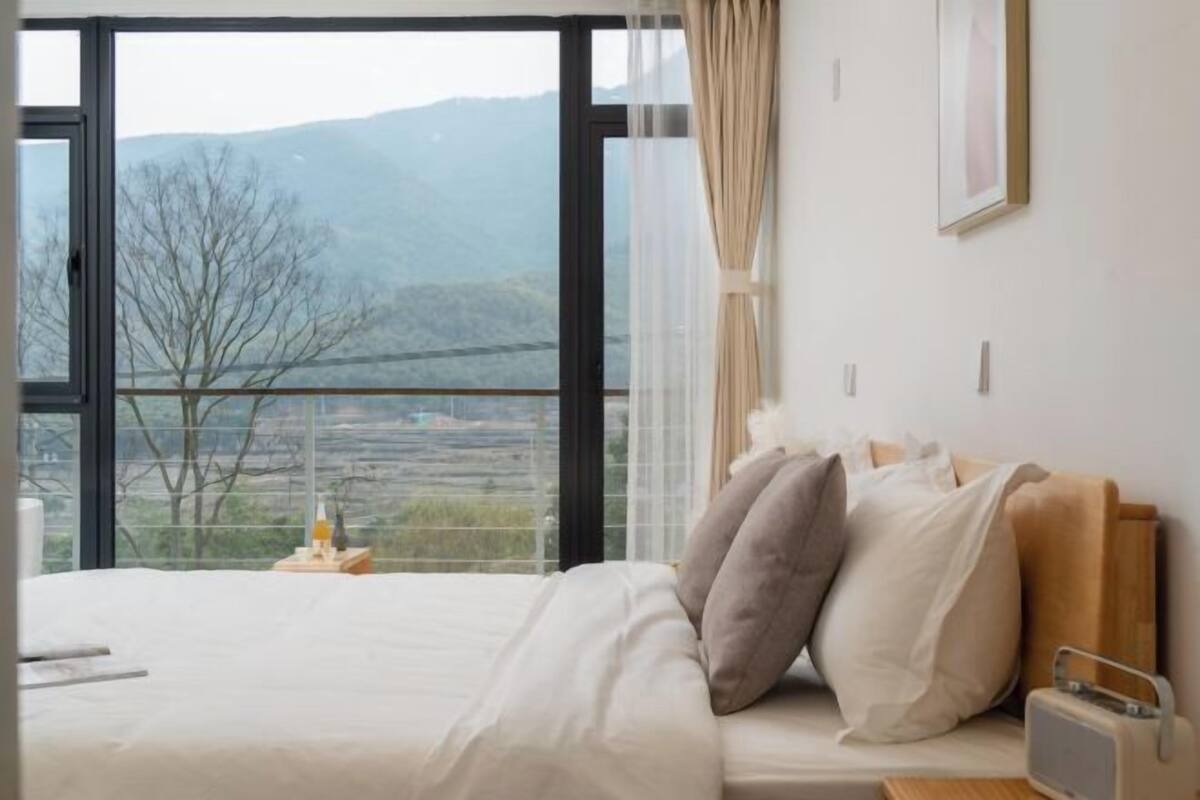 A bedroom with a large window, a bed with pillows, a bedside table with a lamp, and a framed picture on the wall.
