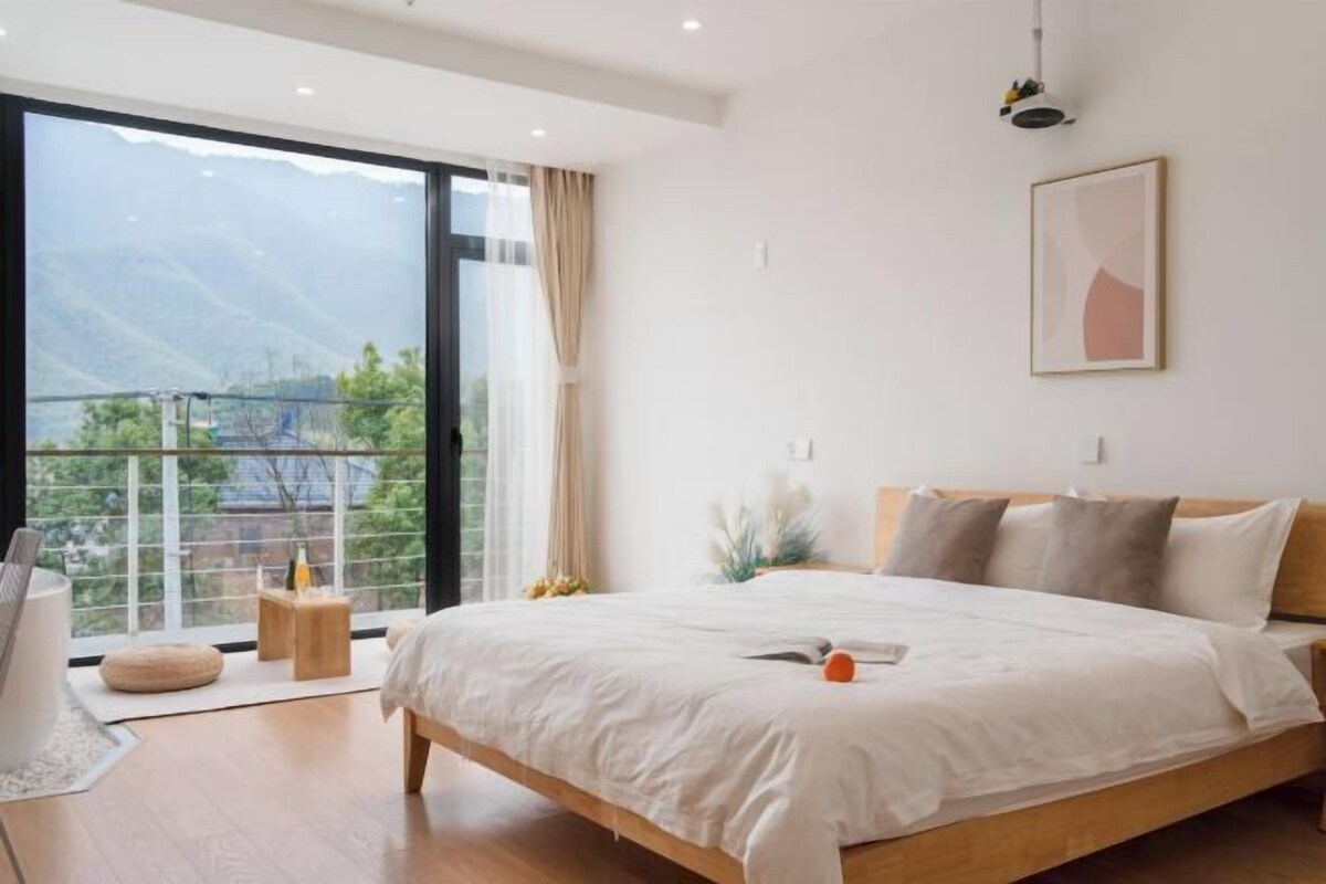 A modern bedroom with a large bed, a bedside table, a window with a view of mountains, and a framed picture on the wall.