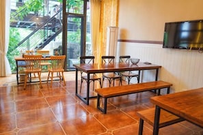 A dining area with wooden tables and chairs, a TV mounted on the wall, and a view of an outdoor staircase. - Yuanshan Hostel (Deqing)