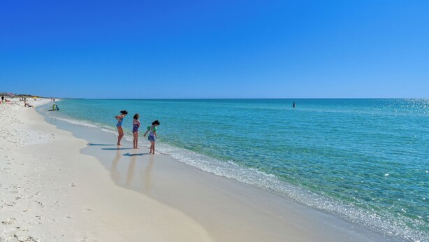 Une plage avec une eau d’un bleu limpide et quelques personnes debout sur le sable.