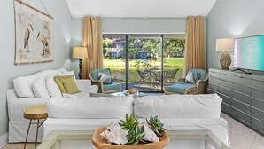 Un salon avec un canapé blanc, deux fauteuils bleus, une table basse et un téléviseur posé sur un meuble.
