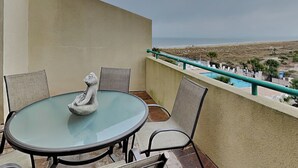 Ein Balkon mit rundem Glastisch und Stühlen, einer dekorativen Steinskulptur sowie Blick auf Strand und Meer.
