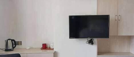 Una habitación con televisión de pantalla plana en la pared, un escritorio con tetera y una silla.