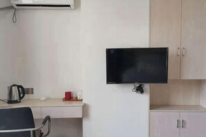 A room with a flat-screen TV mounted on the wall, a desk with a kettle, a chair, and a cabinet.