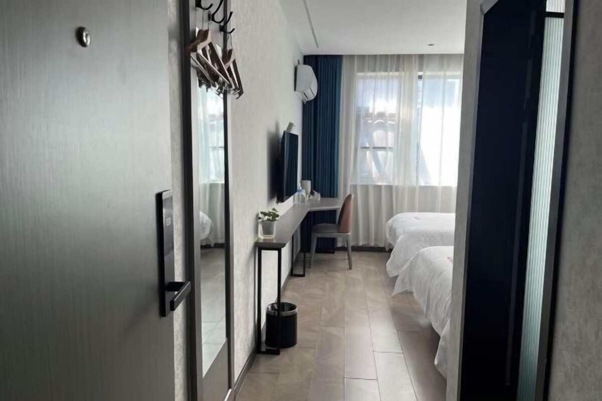 Quarto de hotel com uma cama, uma escrivaninha, uma cadeira, uma mesinha com uma planta, um espelho e uma janela com cortinas.