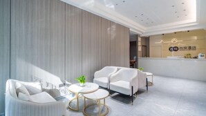 Un hall d’hôtel moderne avec un comptoir d’accueil, un espace salon doté de canapés blancs et de tables rondes, et une grande fenêtre laissant entrer la lumière naturelle.