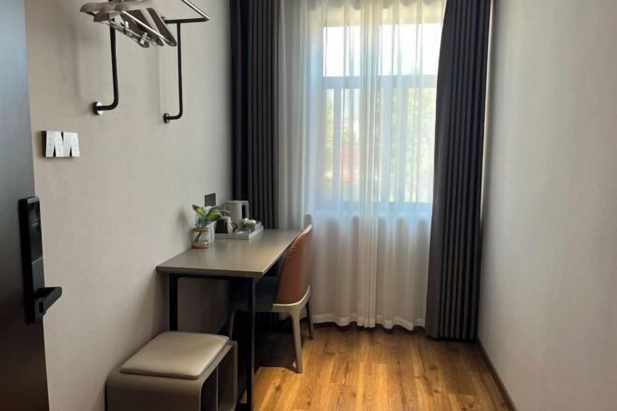 Quarto pequeno e moderno, com piso de madeira, uma escrivaninha com cadeira, um banquinho e uma janela com cortinas.