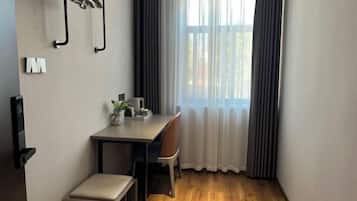 A small, modern room with a wooden floor, a desk with a chair, a small bench, and a window with curtains.