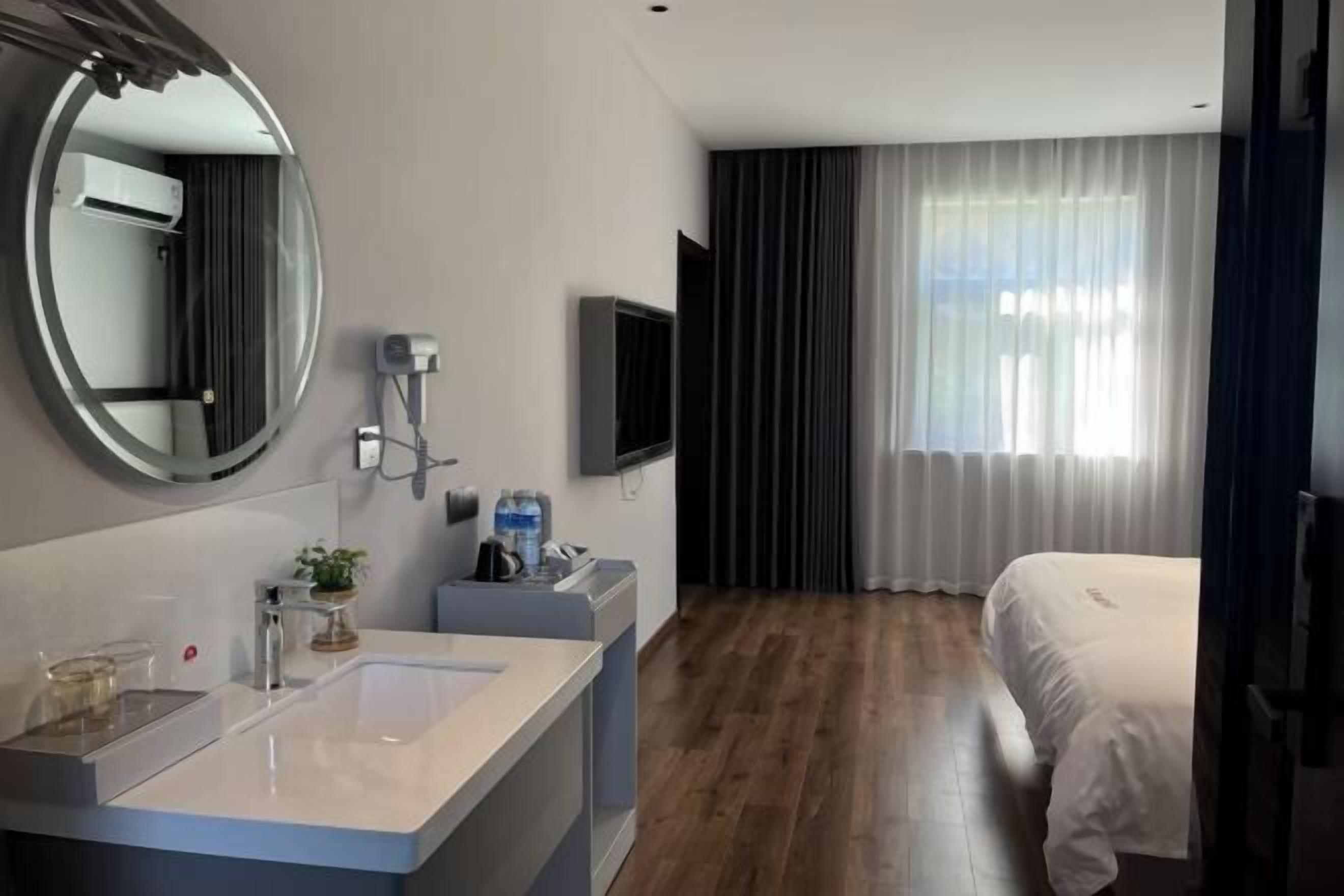 A modern hotel room with a large round mirror, a flat-screen TV, a large bed, and a window with curtains.