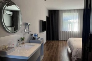 Una habitación de hotel moderna con un espejo redondo grande, un televisor de pantalla plana, una cama grande y un ventanal con cortinas.
