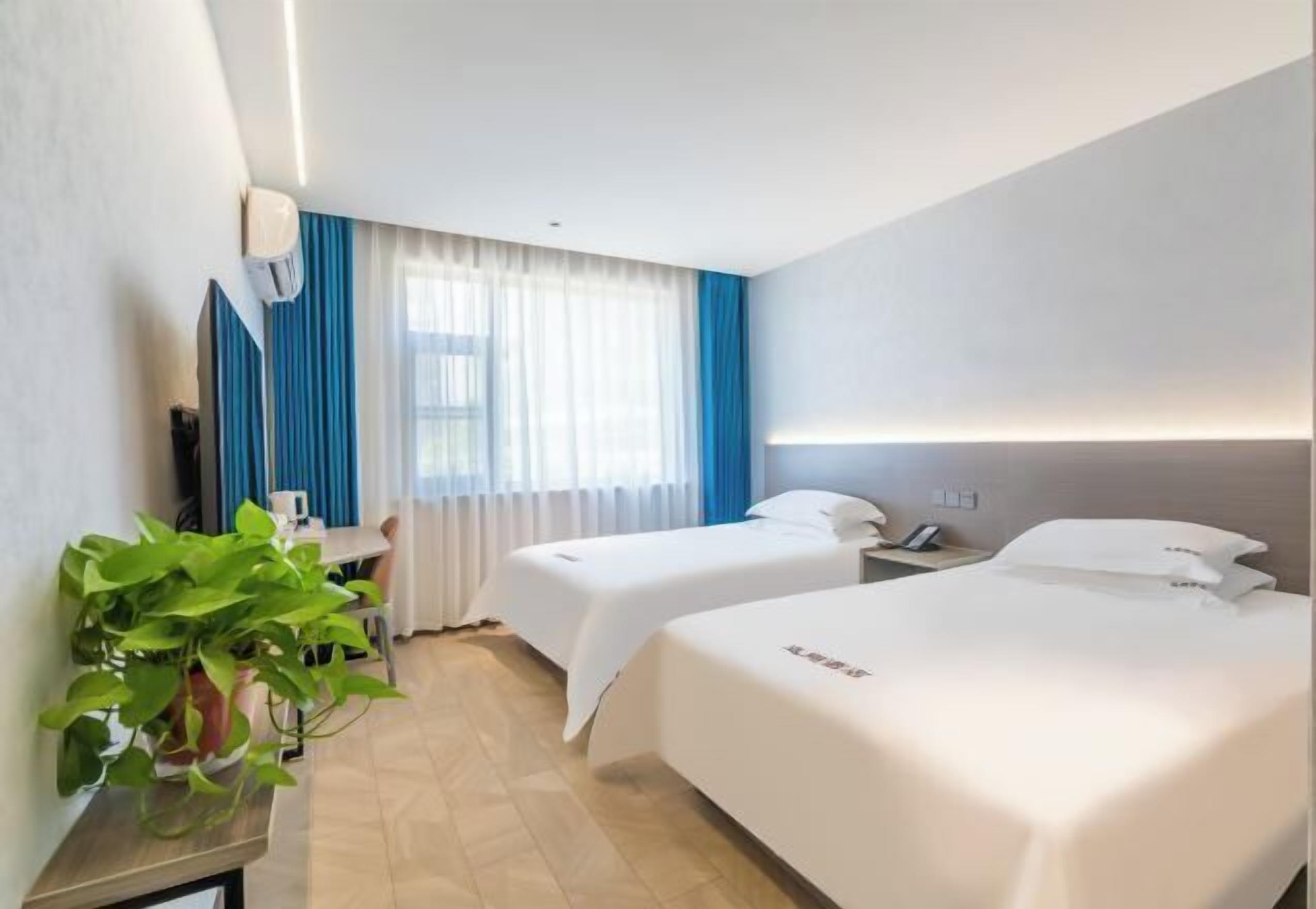 Habitación de hotel con dos camas, un escritorio, una silla, una planta en maceta y una ventana con cortinas.