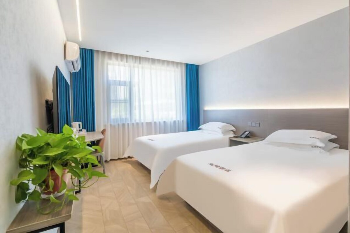 Quarto de hotel com duas camas, uma escrivaninha, uma cadeira, uma planta em vaso e uma janela com cortinas.