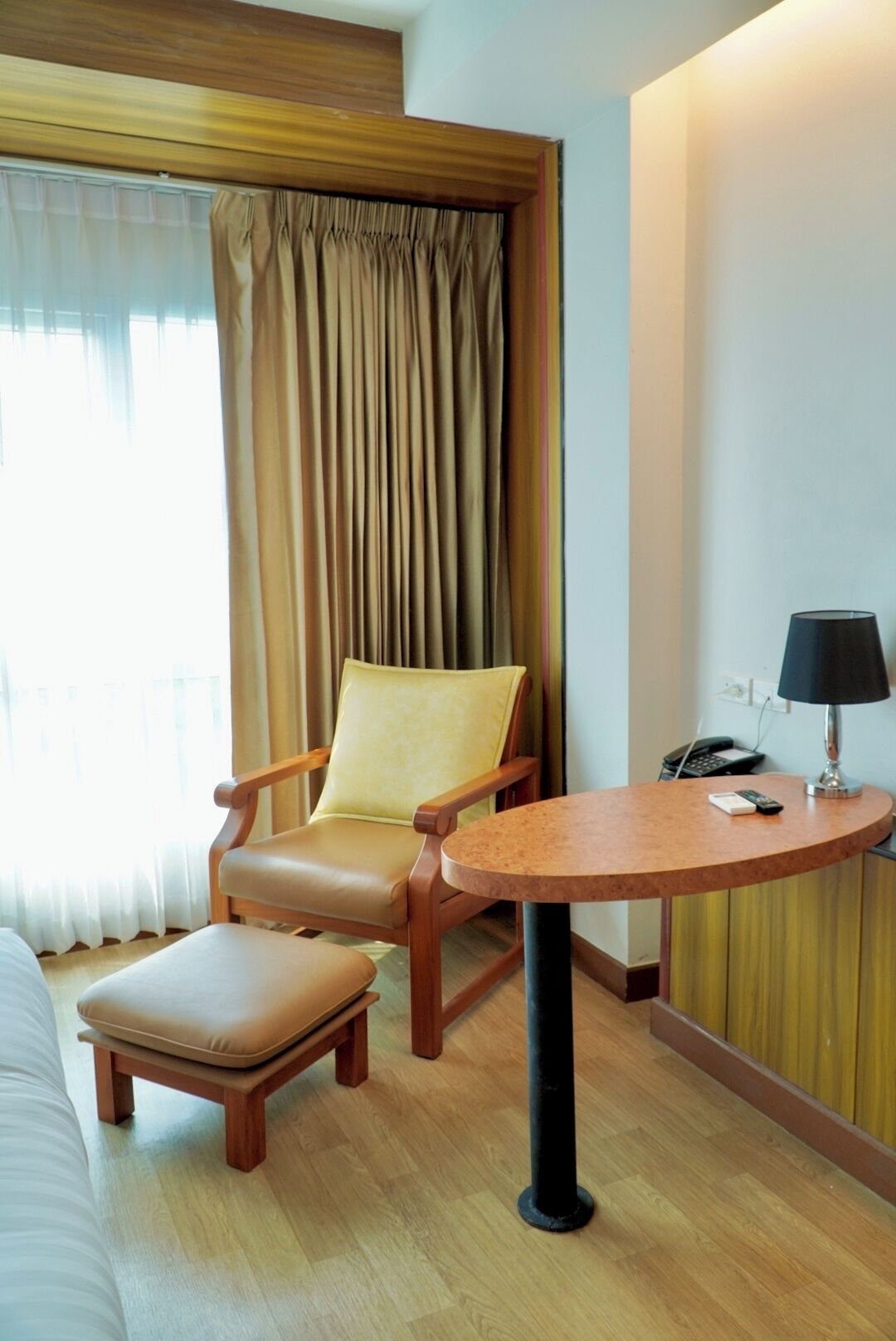 A hotel room with a wooden armchair, a small round table with a lamp, a telephone, and a window with curtains.