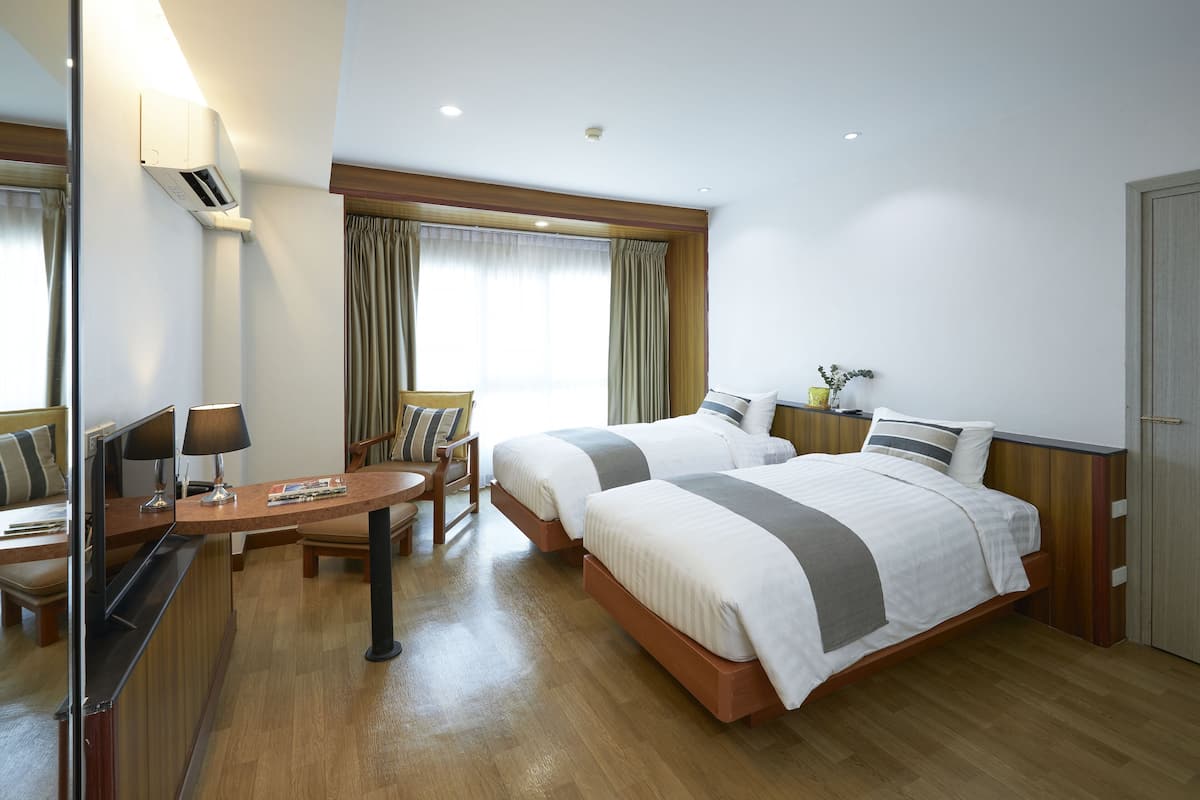 A hotel room with two beds, a desk, a chair, a TV, and a bedside table with a lamp.
