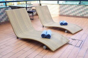 Two wicker lounge chairs with towels, a table, and chairs in a covered outdoor area with a tiled floor.