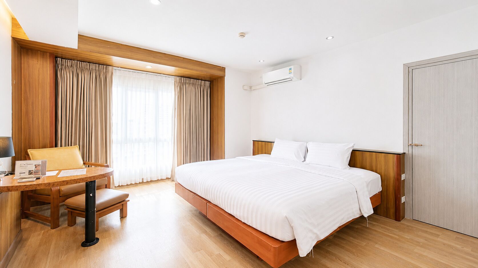 Ein Hotelzimmer mit einem Bett, einem Schreibtisch mit Stuhl, einem Fenster mit Vorhängen und einer Klimaanlage.