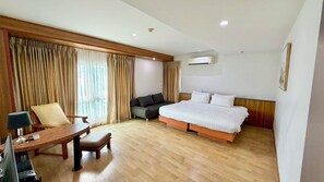 A hotel room with a wooden floor, a bed with white bedding, a sofa, a desk with a lamp, and a window with curtains.