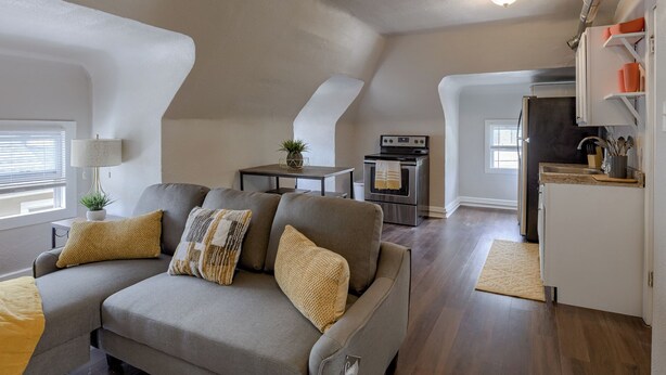 Un appartement-loft confortable avec un canapé gris, une petite table à manger, un coin cuisine et un plafond avec des poutres apparentes.