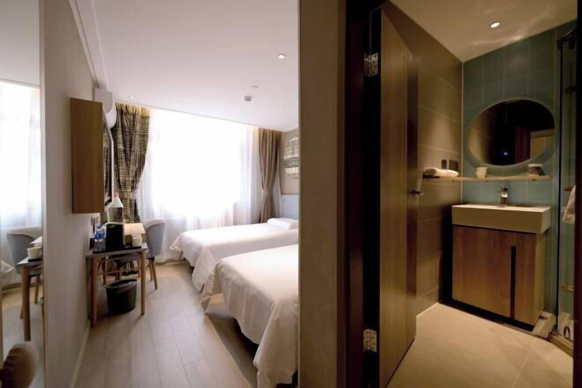 A hotel room with two beds, a desk with a chair, a small table, a mirror, and a bathroom with a sink and mirror.
