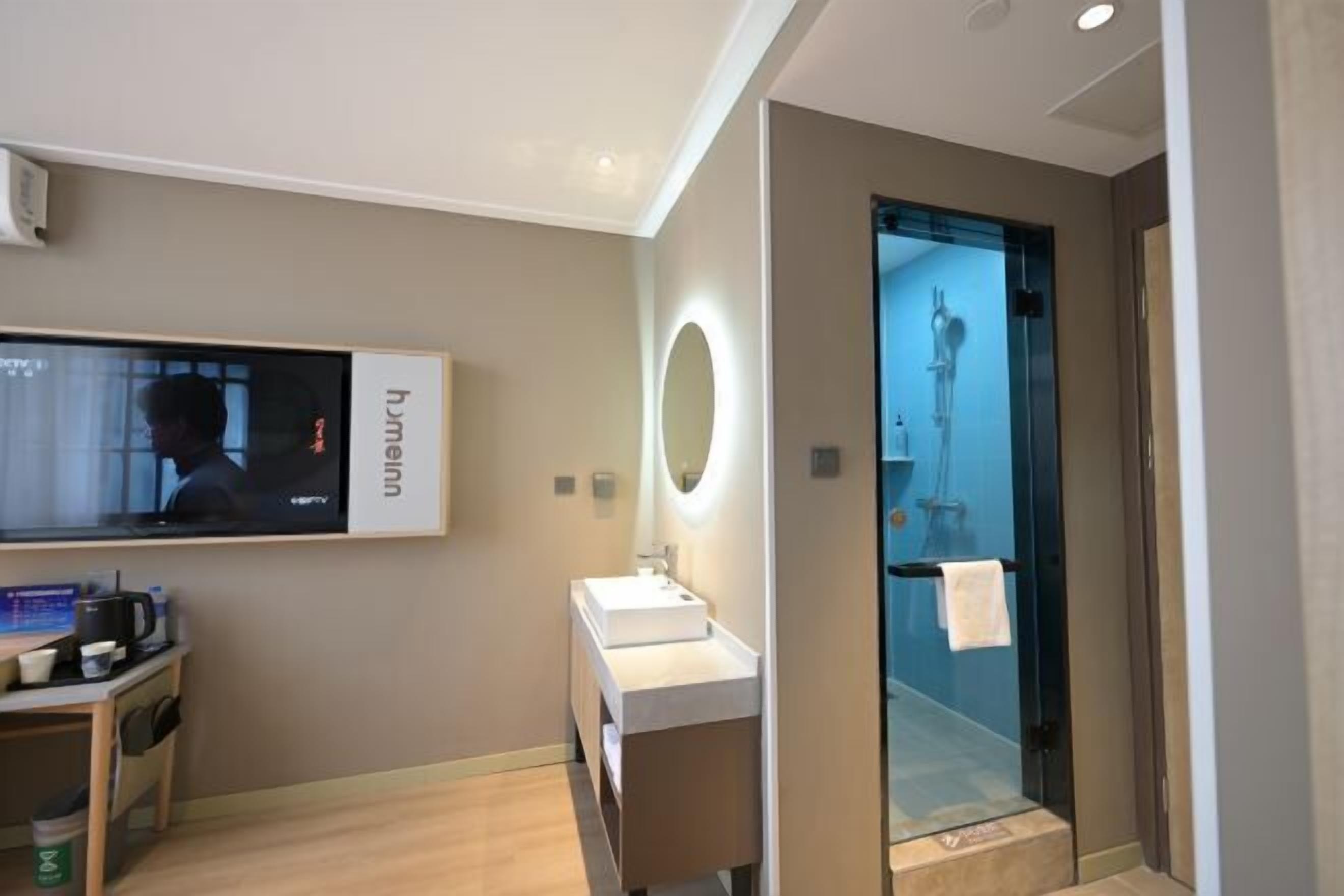 A modern hotel room with a large TV, a bedside table with a coffee maker, a mirror with a light, and a glass shower enclosure.