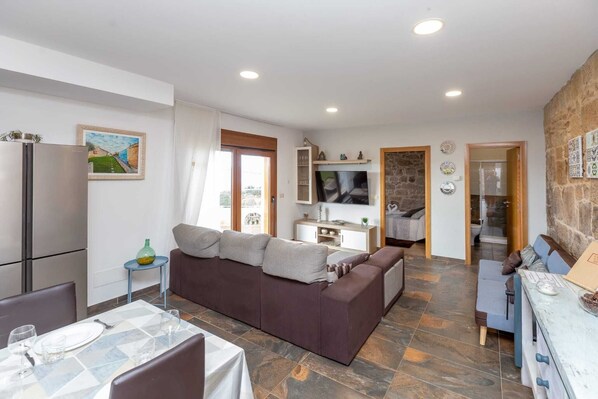 A modern living room with a brown sofa, dining table, and a kitchen area with a TV and stone wall.