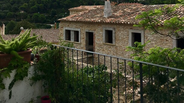Un edificio de piedra con techo de tejas, rodeado de abundante vegetación y un cielo nublado.