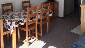 A small, sparsely furnished room with a single table and four chairs, a television on a wooden stand, and a door leading to another room.