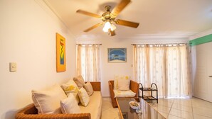 A living room with a sofa, a glass-top coffee table, a ceiling fan, and a view of the outside through curtains. - Garden Condos 40, 1br Apt In Sosua Strip! Guest Ok (Sosúa)