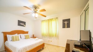 A bedroom with a wooden bed, a ceiling fan, a window with curtains, a desk with a computer and remote controls, and artwork on the walls. - Garden Condos 40, 1br Apt In Sosua Strip! Guest Ok (Sosúa)