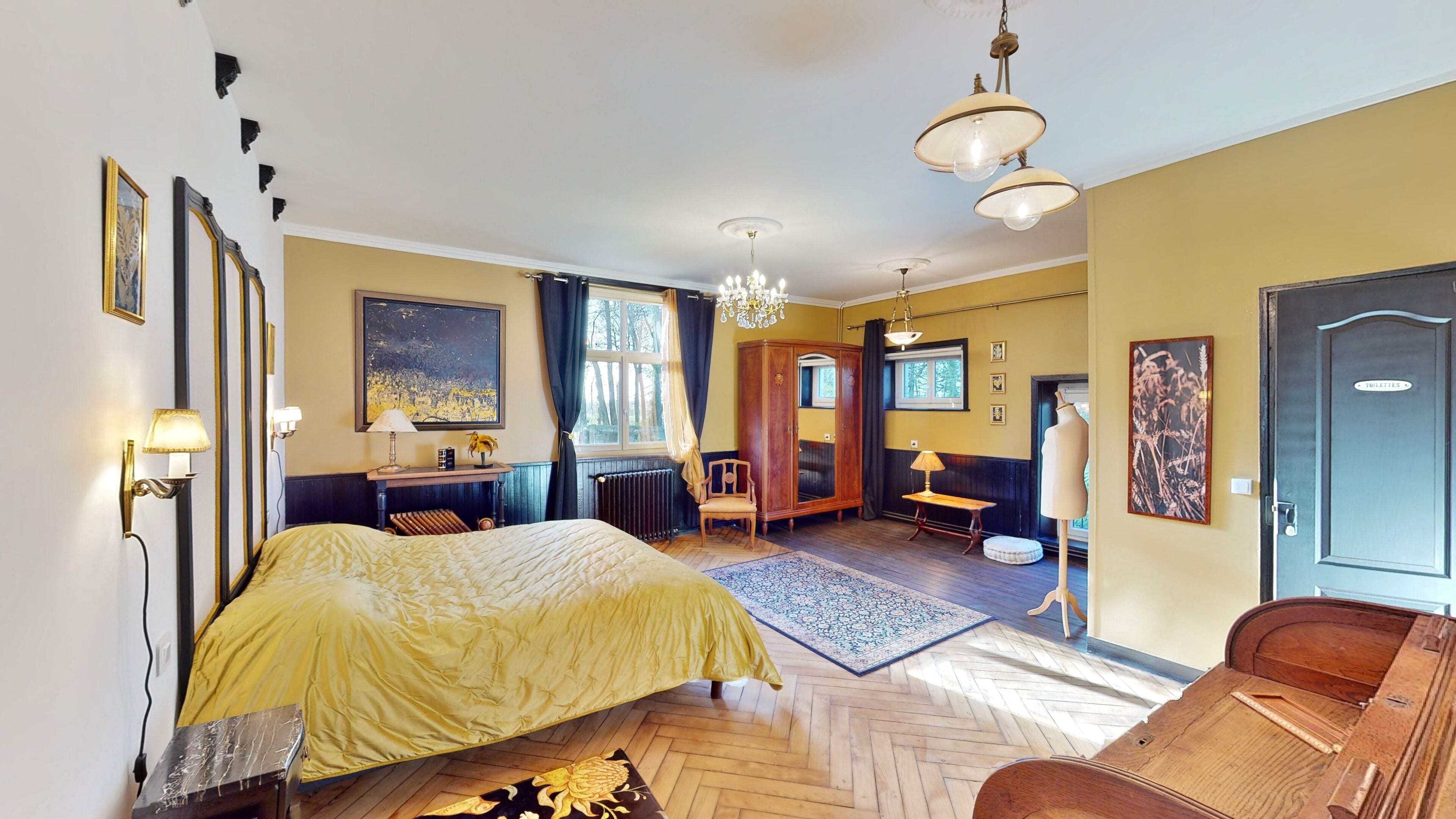 A spacious bedroom with a large bed, a wooden wardrobe, a desk with a chair, a chandelier, and a view of the outdoors.