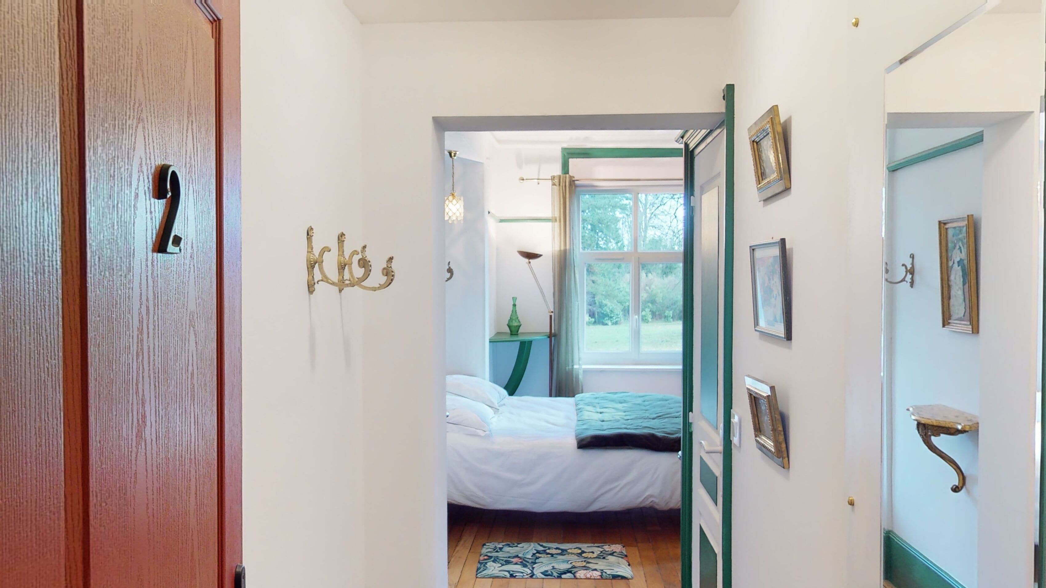 A narrow hallway leading to a bedroom with a bed, a window, and a door with a brass handle.