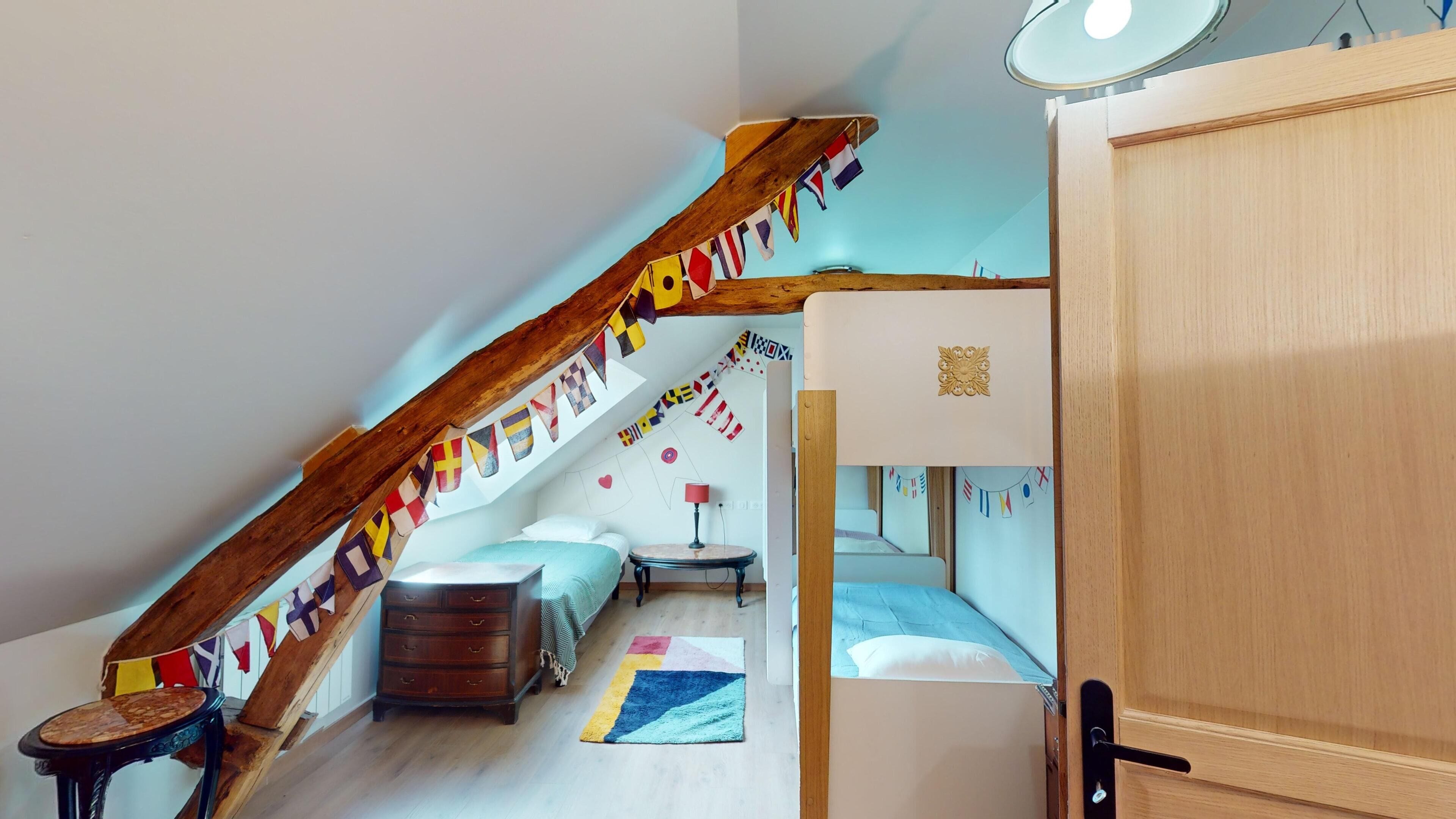 A cozy attic bedroom with a sloped ceiling, wooden beams, a bunk bed, a small table, and a wardrobe.