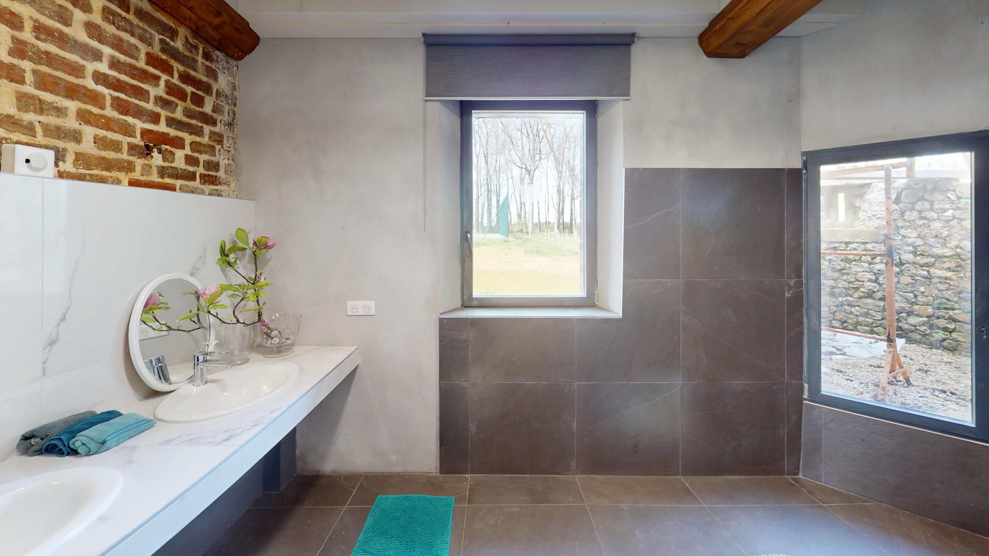 Une salle de bain avec un lavabo en marbre blanc, un miroir, une fenêtre donnant sur la verdure et un espace douche aux carreaux foncés.