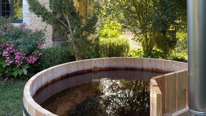 Una bañera de hidromasaje de madera rodeada de exuberante vegetación, con un edificio de piedra al fondo.