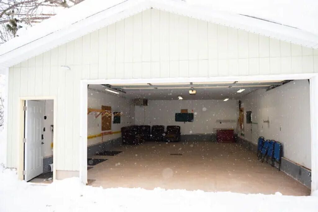 A snow-covered garage with a white exterior, a door leading inside, and a few chairs stacked against the wall.