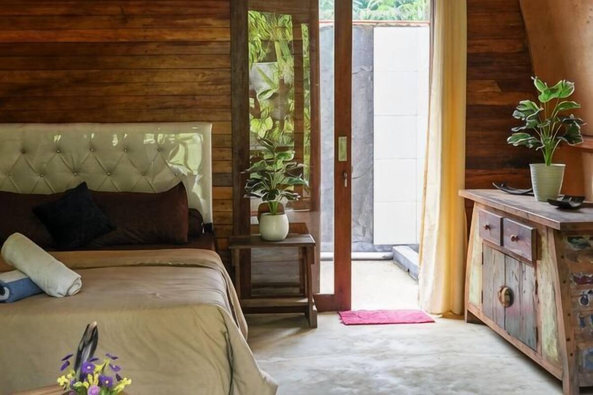 A bedroom with a bed, a wooden headboard, a potted plant, a wooden cabinet, and a door leading outside.