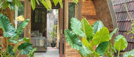 A wooden building with a bed and a small table, surrounded by large green plants.