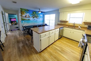 A kitchen with wooden flooring, white cabinets, a granite countertop, a built-in dishwasher, and a mural of a tropical scene.