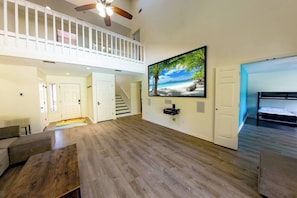 A spacious living room with a high ceiling, wooden flooring, a large TV mounted on the wall, a staircase leading to an upper level, and a bed in the adjacent room.