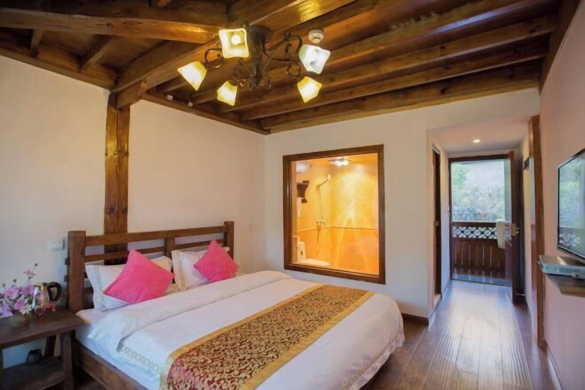 A bedroom with a wooden ceiling, a large bed with white bedding and a patterned throw, a nightstand with a vase of flowers, and a view of a bathroom through a window.