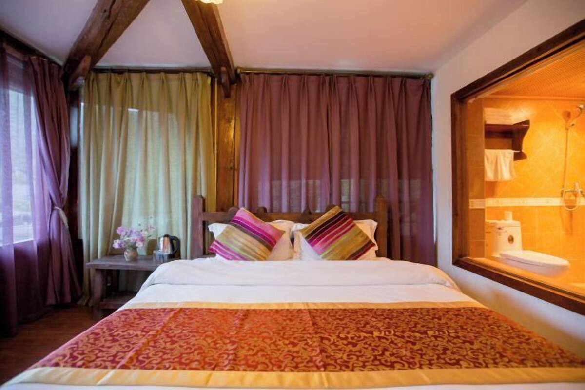 A bedroom with a bed, curtains, a wooden headboard, a nightstand with a lamp, and a window with a view of a bathroom.