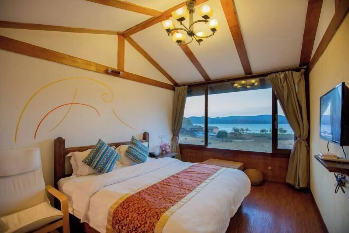 A bedroom with a large bed, wooden beams, a chandelier, a TV, and a view of the landscape.